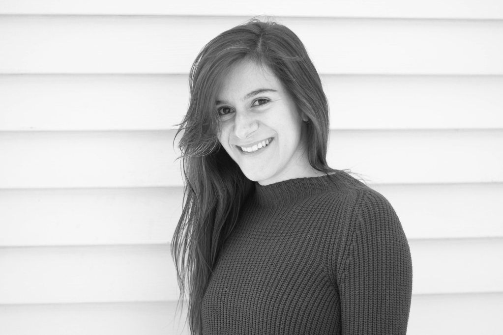 A woman, author Nicole Haldoupis, stands in front of a white, planked wall for a portrait.